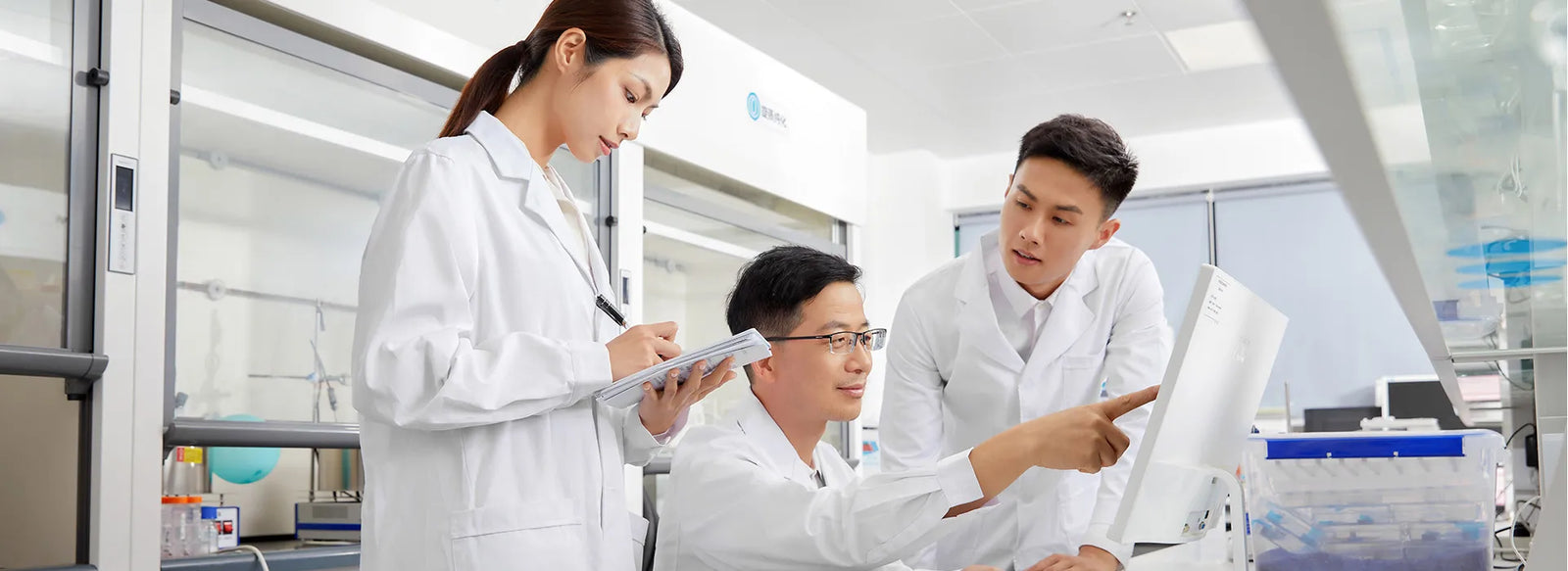 Three lab professionals in white coats examine a computer screen. One writes on a clipboard. Lab equipment in background. Keywords: Sibionics, blood glucose monitor, lab, professionals.
